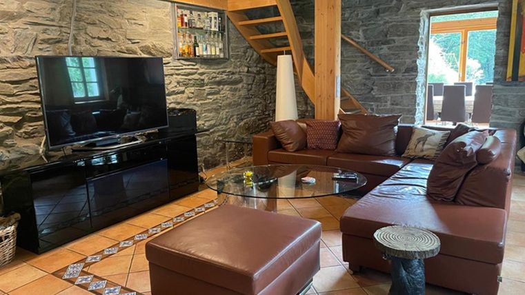 Een gezellige woonkamer met een bruine bank en een glazen tafel. De ruimte heeft stenen muren en grote ramen die veel daglicht binnenlaten.