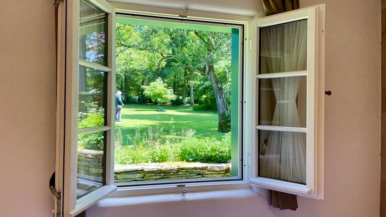 Ein offenes Fenster mit Blick auf einen grünen Garten. Im Hintergrund sind Bäume und eine Person zu sehen.