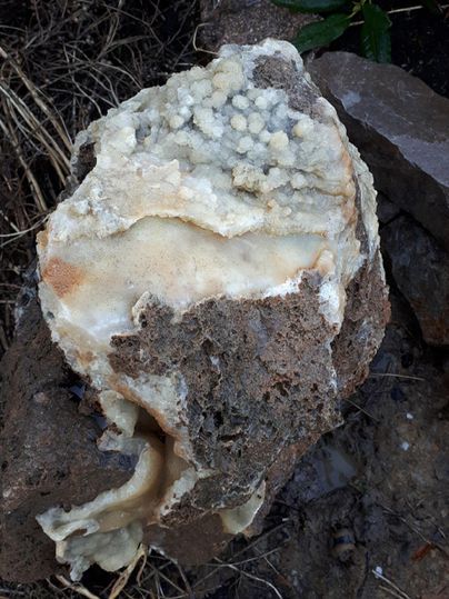 Ein Stück Gestein mit einer rauen, braunen Oberfläche, das von kristallinen Strukturen überzogen ist. Die Kristalle haben eine helle, fast milchige Farbe.