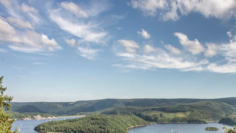 Un lac pittoresque entouré de collines verdoyantes et de rives boisées. Le ciel est bleu avec quelques nuages.