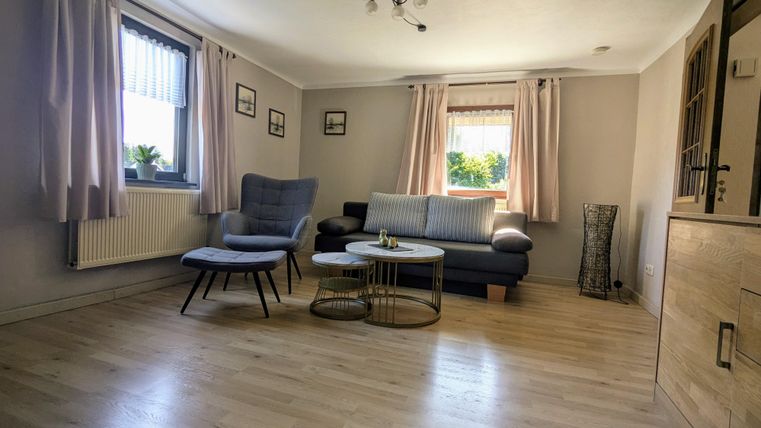 Ein helles Wohnzimmer mit Holzfußboden und großen Fenstern. Es gibt gemütliche Möbel, darunter einen Sessel und ein Sofa.