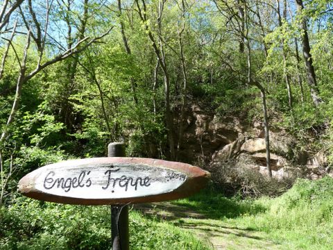 A sign with the inscription "Engel's Tropp" stands in front of a wooded area. The surroundings are green and lush, with trees and a rock in the background.