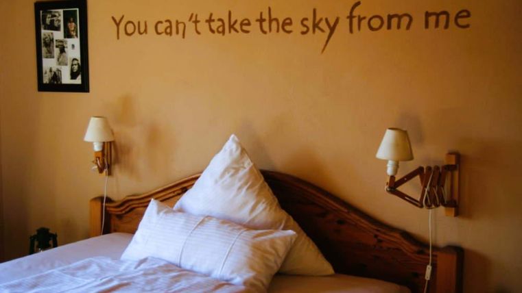 Ein gemütliches Schlafzimmer mit einem großen Bett und dekorativen Kissen. An der Wand steht der Spruch: „You can't take the sky from me“.