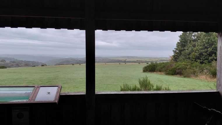Ein Blick aus einem Fenster auf eine weite, grüne Landschaft mit leicht bewölktem Himmel. Im Vordergrund steht ein Informationsschild.