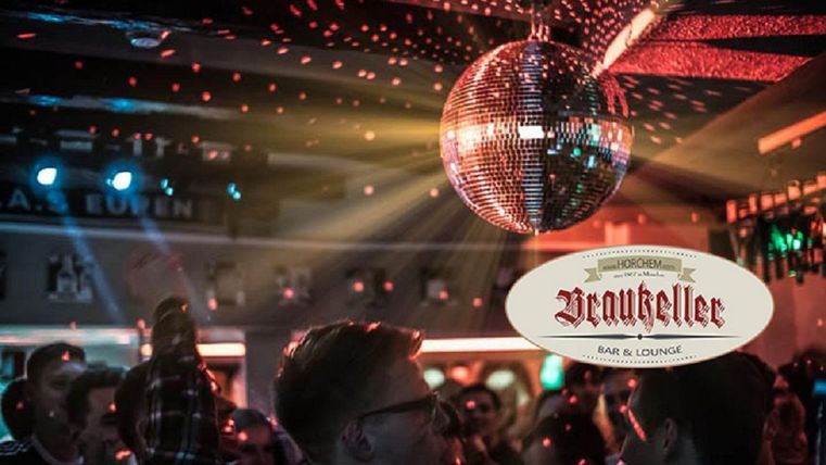 Eine lebhafte Bar mit tanzenden Menschen und einer glitzernden Discokugel. Bunte Lichter schaffen eine festliche Atmosphäre.