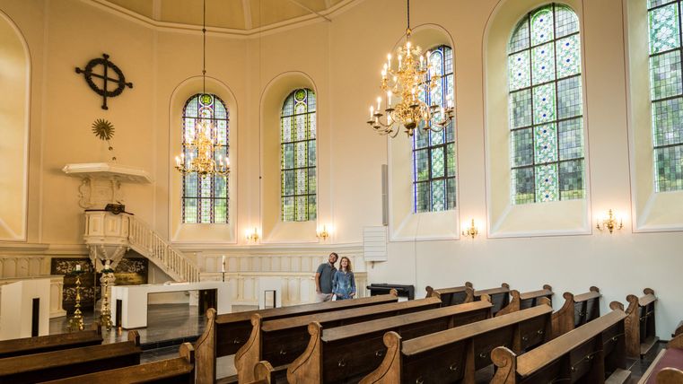 Une église tranquille avec des rangées de bancs en bois et des fenêtres colorées. Au premier plan, deux personnes observent l'espace.