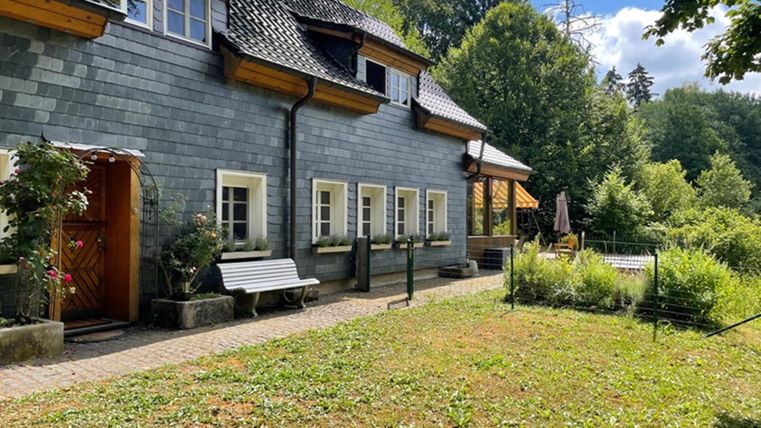Een charmant, grijs huis met een mooie tuin. Op de achtergrond zijn bomen en een zonnig landschap te zien.