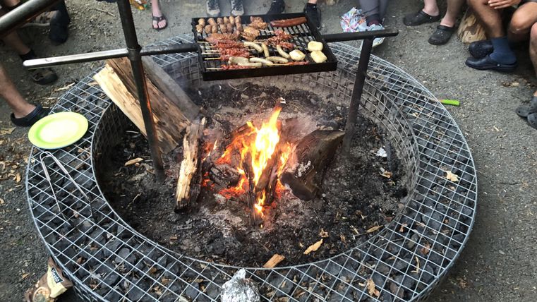 Ein Lagerfeuer mit einem Grill, auf dem verschiedene Speisen zubereitet werden. Im Hintergrund sind Menschen versammelt und genießen die Campingatmosphäre.
