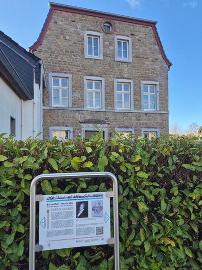 Un bâtiment historique doté de nombreuses fenêtres et d'un toit rouge. Au premier plan se trouve un panneau d'information qui donne plus de détails sur l'histoire de la maison.