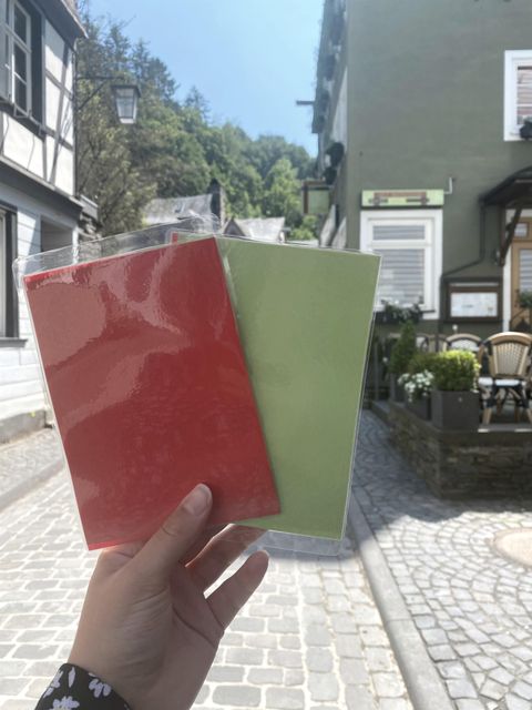Eine Hand hält drei farbige Notizhefte, rot und grün, vor einer malerischen Gasse. Im Hintergrund sind historische Gebäude und eine gemütliche Terrasse zu sehen.