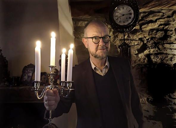 Ein Mann in einem dunklen Raum hält einen Kerzenleuchter mit leuchtenden Kerzen. Im Hintergrund sind eine Uhr und Steinwände sichtbar.
