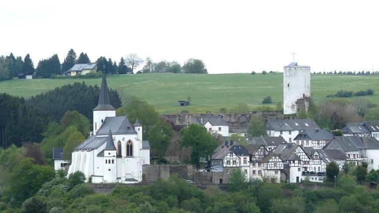 Un village pittoresque avec une église et des maisons traditionnelles. À l'arrière-plan, on aperçoit des collines verdoyantes et un belvédère.
