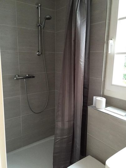Une douche moderne dans une salle de bain lumineuse. Les murs sont recouverts de carreaux gris et un rideau de douche assure l'intimité.