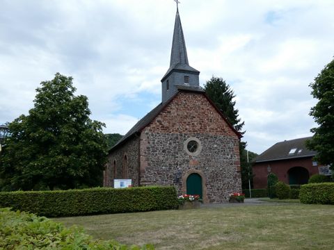 Eine kleine, historische Kirche mit einem spitzen Turm und umgeben von grünen Bäumen. Der Garten ist gepflegt und die Atmosphäre ist ruhig und einladend.