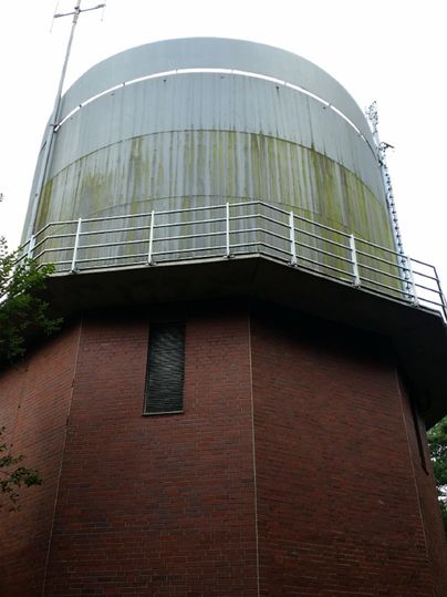 Ein Wasserhochbehälter mit einem runden, metallenen Oberteil. Der untere Teil besteht aus rotem Backstein und ist von Bäumen umgeben.