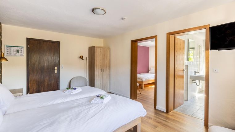 Ein helles Zimmer mit zwei Einzelbetten und modernen Möbeln. Im Hintergrund sieht man eine offene Tür zu einem Badezimmer und einen weiteren Schlafraum.