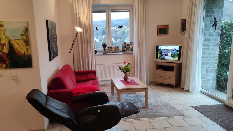 Ein gemütliches Wohnzimmer mit einem roten Sofa und einem relaxenden Sessel. Ein Tisch mit einer Blumenvase und ein Fernseher sind ebenfalls sichtbar.