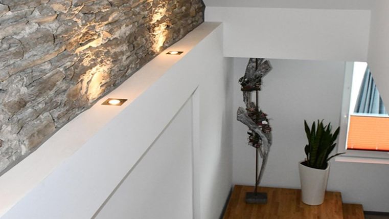 Un espace d'escalier moderne avec un mur en pierre et des lumières élégantes. À côté de l'escalier se trouve une plante dans un pot blanc et une sculpture décorative.