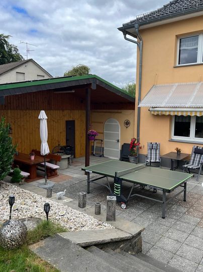 Ein gemütlicher Patio mit einem Tischtennisplatz und einem Holzpavillon. Der Bereich ist von Pflanzen und einem hellen Haus umgeben.