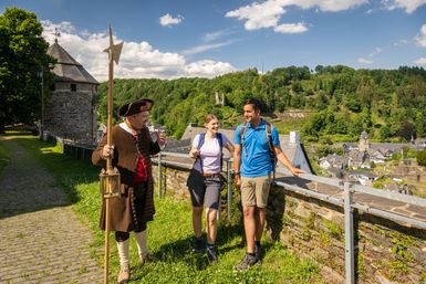 Auf dem Bild sind drei Personen zu sehen, zwei davon sind Teilnehmer einer geführten Tour und die dritte ist der Tourguide verkleidet als Stadtwächter. Im Hintergrund ist die historische Altstadt von Monschau.