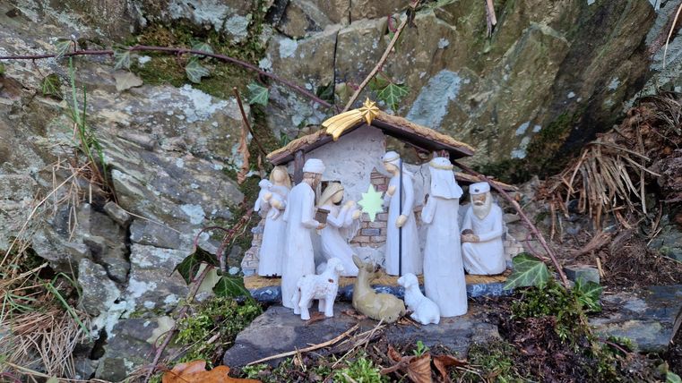 Eine kleine Krippe mit Figuren, die die Geburt Jesu darstellen, steht vor einer Felsenwand. Umgeben von Moos und Laub vermittelt die Szene eine besinnliche Atmosphäre.