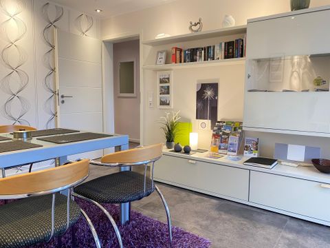 Een moderne woonkamer met een eettafel en stoelen. Aan de muur hangt een rek met boeken en decoratieve elementen.