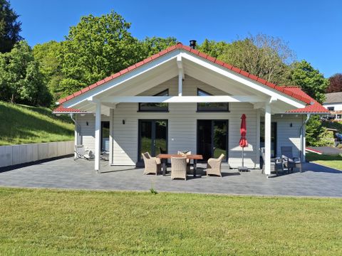 Ein modernes Haus mit rotem Dach und Veranda. Der Garten ist grün und gut gepflegt, umgeben von Bäumen.