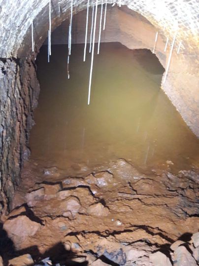 Eine dunkle, unterirdische Höhle mit steinigen Wänden und stagnierendem Wasser. Es hängen stalaktitenartige Formen von der Decke.