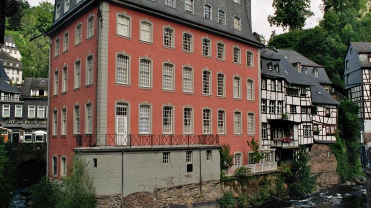 Ein historisches Gebäude mit roter Fassade steht am Ufer eines klaren Baches. Im Hintergrund sind Fachwerkhäuser und grüne Bäume zu sehen.