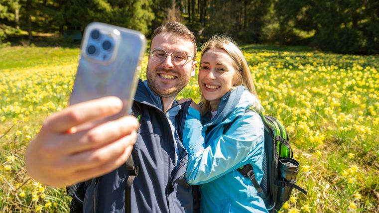 Ein Paar macht ein Selfie vor einer Wiese voller gelber Narzissen, umgeben von Bäumen.