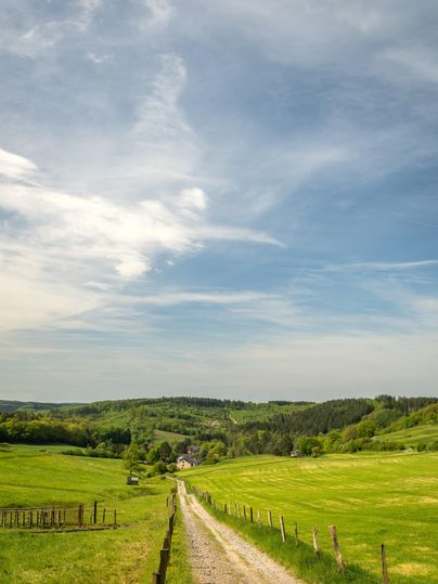 A picturesque landscape with a gravel path leading through green meadows. The sky is blue with some clouds.