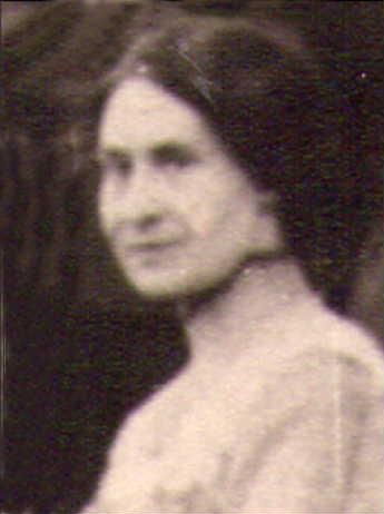 A blurry portrait of an older woman with a serious expression. She is wearing a simple dress and has long, dark hair.