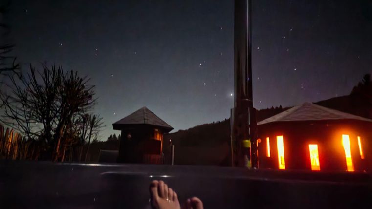 Ein entspannendes Bad im Freien unter einem klaren Sternenhimmel. Im Hintergrund sind zwei beleuchtete Gebäude und Bäume sichtbar.