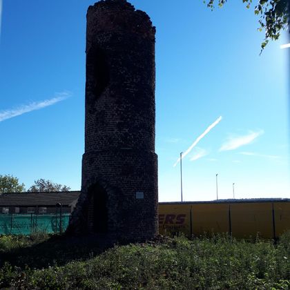 Ein alter, gebrochener Turm aus Stein steht in einer grünen Wiese. Im Hintergrund sind ein blauer Himmel und einige Wolken zu sehen.