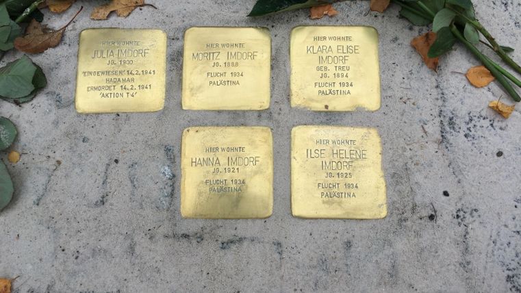 Six brass commemorative plaques lie on the ground, surrounded by rose petals. Each plaque bears the name and biographical details of individuals.