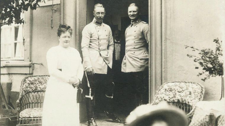 Drei Personen stehen vor einem Eingang: zwei Männer in historischen Uniformen und eine Frau in einem weißen Kleid. Im Hintergrund sieht man einige Sitzmöbel und eine blühende Pflanze.