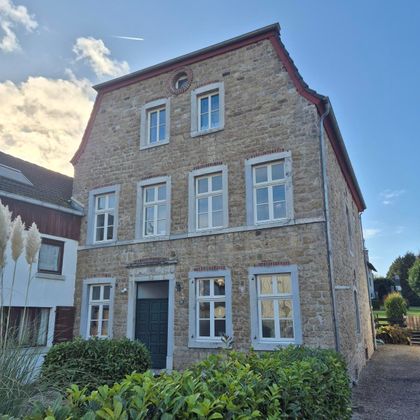 Une maison historique en pierre aux fenêtres blanches et à l'entrée verdoyante. Le ciel est dégagé, avec quelques nuages.