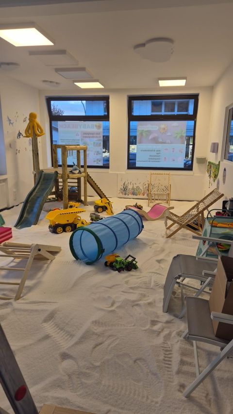 Ein Spielzimmer mit Sandboden und verschiedenen Spielgeräten. Spielzeug wie Autos und ein Tunnel sind ebenfalls vorhanden.