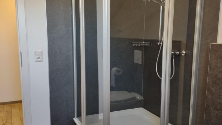 Une cabine de douche moderne avec des parois en verre clair se trouve dans une salle de bains lumineuse. Le sol est recouvert de parquet, et il y a une porte qui mène à une autre pièce.