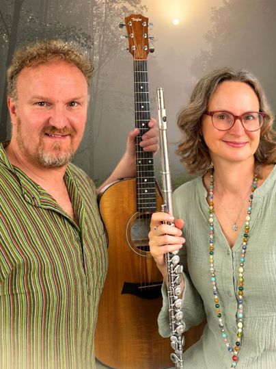 Un homme avec une guitare et une femme avec une flûte se tiennent ensemble devant un arrière-plan de forêt pittoresque. Tous deux sourient amicalement et portent des vêtements décontractés.