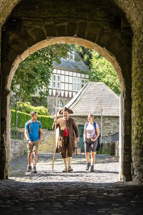 Drei Personen, darunter ein Stadtwächter in historischer Kleidung, gehen gemeinsam durch ein historisches Tor der Burg Monschau.