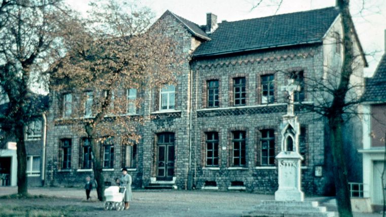 An old building with red bricks and large windows stands in the foreground. In the background, you can see some trees and a statue.