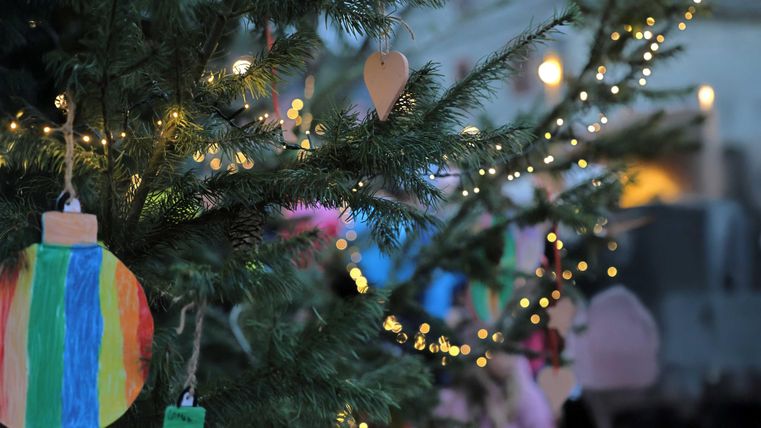 Ein geschmückter Weihnachtsbaum mit bunten Ornamenten und Lichtern. Im Hintergrund sind unscharfe Silhouetten von Menschen zu sehen.