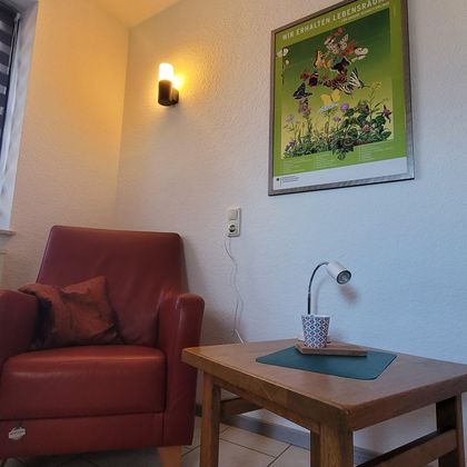 Ein gemütlicher Raum mit einem roten Sessel, einem kleinen Holztisch und einer Lampe. An der Wand hängt ein buntes Poster.