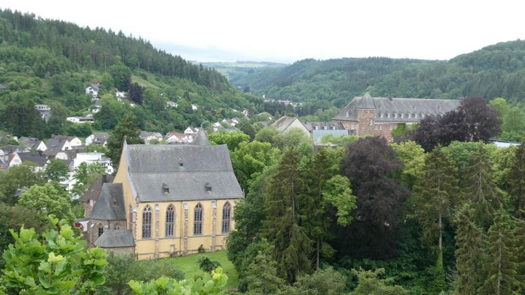 Eine malerische Landschaft mit einer Stadt, umgeben von grünen Hügeln. Im Vordergrund ist ein historisches Gebäude sichtbar, das von Bäumen umgeben ist.