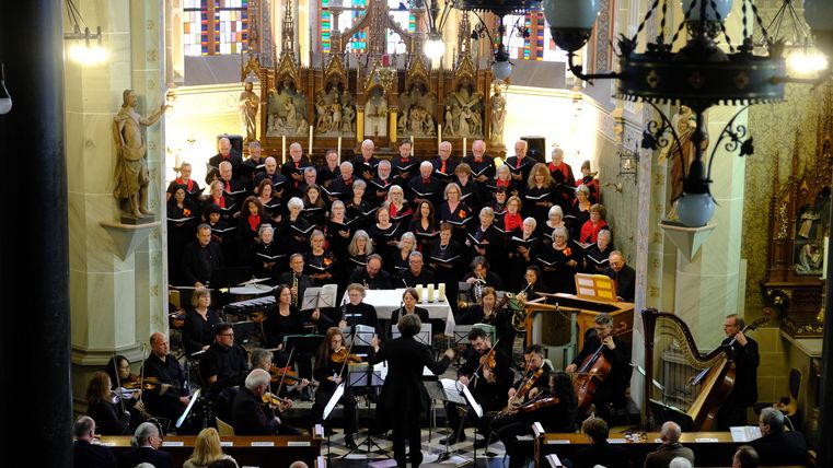 Ein großer Chor singt in einer Kirche, umgeben von musikalischen Begleitern. Die farbenfrohen Glasfenster sorgen für eine festliche Atmosphäre.