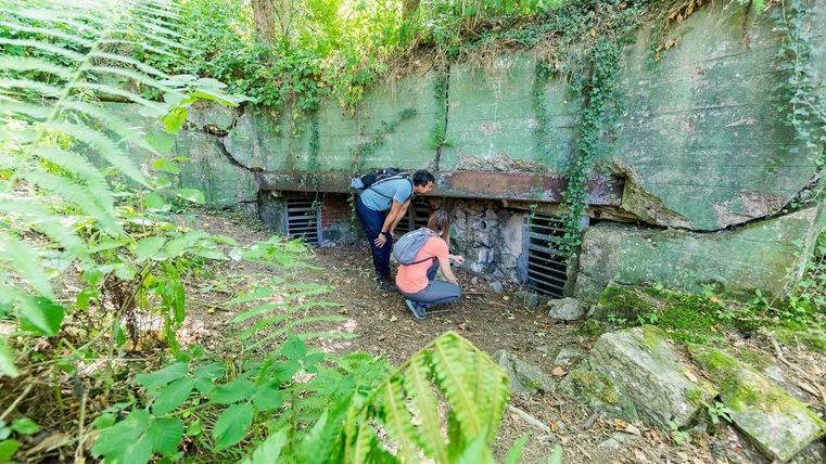 Zwei Personen erkunden einen alten Bunker, der von Pflanzen umgeben ist. Die Umgebung ist üppig und grün, was dem Bild eine geheimnisvolle Atmosphäre verleiht.