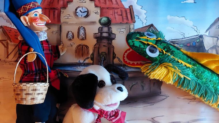 Eine bunte Puppenspiel-Szene mit einer Marionette, einem Stoffhund und einem grünen Drachen. Im Hintergrund ist ein malerischer Brunnen und ein Gebäude zu sehen.