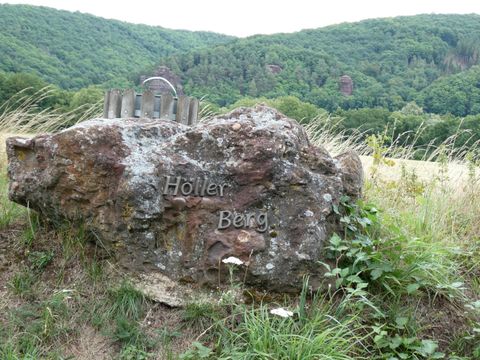 A large rock with the inscription "Holler Berg" stands in a green landscape. In the background, there are gentle hills and trees.