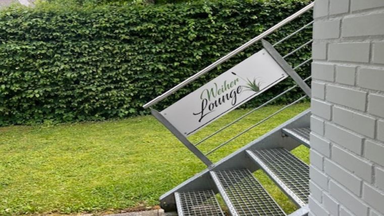 Eine Außenansicht mit einer Metalltreppe und einem Schild, das auf die "Welker Lounge" hinweist. Im Hintergrund sind grüne Sträucher und ein gepflegter Rasen zu sehen.